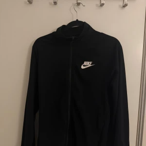 Svart Nike vindjacka med dragkedja - Svart dress från Nike med klassisk vit logga på bröstet. Jackan har hel dragkedja framtill, hög krage och långa ärmar. Perfekt för träning eller chill dagar. Materialet är lätt och känns sportigt.