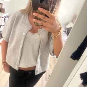 Ljusgrå stickad kofta med kort ärm - Säljer en ljusgrå stickad kofta med korta ärmar och rund hals. Koftan har ribbad struktur och knäpps med vita knappar framtill. Perfekt att slänga över en topp för en chill och snygg look. Passar dig som gillar enkel och stilren design.