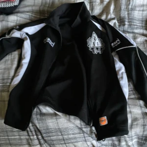 Svartvit träningsväst från ZENI - Sportig svart väst med vita paneler från ZENI losport, prydd med ÖIF-logga på bröstet. Västen har dragkedja framtill och hög krage, perfekt för träning eller lagaktiviteter. Materialet är troligtvis polyester och designen är klassiskt atletisk.