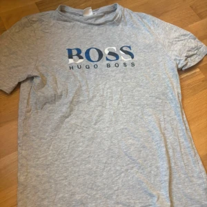 Grå Hugo Boss t-shirt - Snygg grå t-shirt från Hugo Boss med stort BOSS-tryck i blått och vitt på bröstet. Klassisk rund hals och korta ärmar. Mjuk och skön bomull som passar perfekt till jeans eller shorts. Enkelt stilren och lätt att matcha.