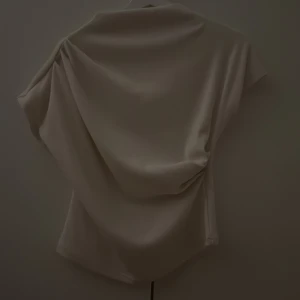 Beige omlott blus i satin - Säljer en beige blus snygg omlott-detalj på ena sidorna. Toppen har korta ärmar och ett mjukt,  tyg som ger en lyxig känsla. Perfekt för dig som gillar stilrena och trendiga plagg med en twist.