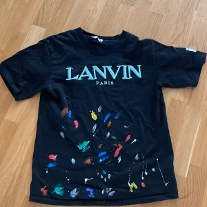 Lanvin x gallery dept - Svart t-shirt från Lanvin Paris med coolt tryck i blått och vitt framtill. Unik design med färgstänk i rött, blått, grönt, gult och orange på framsidan. Kortärmad modell i mjuk bomull. Patch på ärmen med texten 'Art on Display Courtesy of Josué Thomas'. Perfekt för dig som gillar streetstyle.
