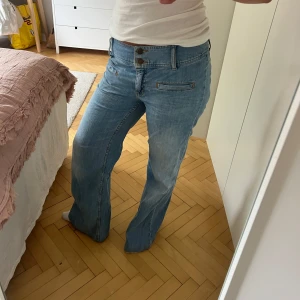 Blå bootcut jeans med dubbla knappar - Säljer ett par ljusblå jeans med bootcut passform och dubbla knappar i midjan. Jeansen har raka ben, klassiska fickor fram och bak samt snygga sömmar. Perfekta för dig som gillar en avslappnad och trendig stil.
