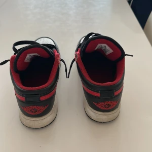 Nike Air Jordan 1 Low svart/röd/vit - Nike Air Jordan 1 Low sneakers med svart och vitt skinn, röda detaljer och röd insida. Klassisk Jordan-logga på hälen och Jumpman på tungan. Perforerad tå och svarta snören ger en clean och sportig vibe. Perfekt för dig som vill sticka ut med ikoniska sneakers.