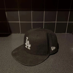 Svart LA-keps från New Era 7 3/8 - Snygg svart keps från New Era med LA-logga framtill i vitt. Klassisk 59FIFTY-modell med böjd skärm och broderade detaljer. Tillverkad i polyester och bomull, perfekt för dig som gillar streetwear och sportig stil.