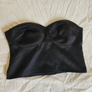 Silkig svart bustier-topp - Korsettliknakde bustiertopp från Gina Tricot, ryschad baksida. Har egentligen löstagbara axelband, men jag har tappat bort det ena tyvärr :( Gummilinningen i bysten är lite sliten, men annars i bra skick