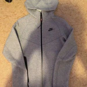 Grå hoodie från Nike med dragkedja - Säljer en snygg grå hoodie från Nike med hel dragkedja och huva. Tröjan har lång ärm och en stilren design med Nike-logga på bröstet. Perfekt för chill eller träning, och den är gjord i mjukt bomullsmaterial för extra komfort.