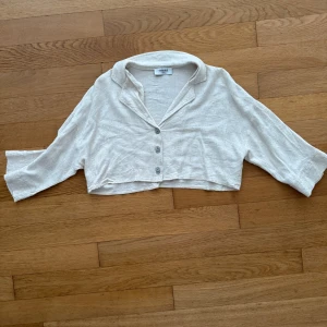 Beige croppad linnetopp från chiquelle - Superfin croppad topp i beige linne. Modellen har tre knappar framtill, krage och vida trekvartsärmar. Perfekt för en chill och avslappnad look med en touch av vintagekänsla.