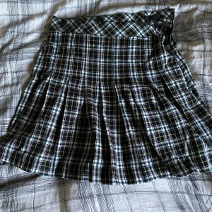 Rutig kortkjol från Lindex - Supersnygg rutig kortkjol från Lindex i storlek 146/152. Kjolen har klassiskt rutmönster i svart, vit, grön och gul med snygga veck och knäppning i midjan. Perfekt för dig som gillar preppy stil och vill sticka ut lite extra.