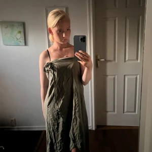 Olivgrön omlottklänning med knyt - Säljer en olivgrön omlottklänning med snygg knytning vid axeln. Klänningen är lång och har en loose passform med ärmlös design. Materialet känns lätt och luftigt, perfekt för varma dagar. Enkel och stilren look som är lätt att matcha med accessoarer.