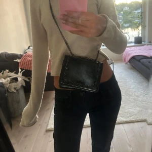 Svart axelväska med kedjedetalj - Snygg svart axelväska i skinn med cool kedja runt kanten och metallkedja som axelrem. Väskan är fyrkantig och har en stilren look som passar till många outfits. Perfekt storlek för mobilen och det viktigaste. Kedjedetaljen ger en edgy vibe.
