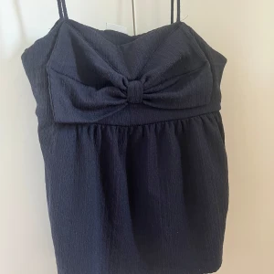 Svart linnetopp med rosett - Supersöt svart linnetopp från Kappahl med smala axelband och en stor rosett framtill. Toppen har en lätt rynkad nederdel och är tillverkad i ett strukturerat tyg som ger en cool känsla. Perfekt för att lyfta vilken outfit som helst!