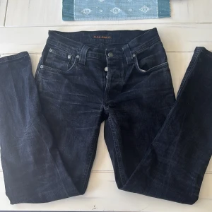 Svarta jeans från Nudie Jeans - Snygga svarta jeans från Nudie Jeans med klassisk femficksdesign och diskreta slitningar. Jeansen har raka ben och normal passform, tillverkade i mjukt denimtyg. Perfekta för dig som gillar stilrena och tidlösa jeans med en cool vibe. W29 L32 grim trim