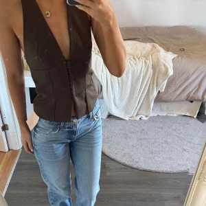 Brun korsett-topp med djup v-ringning - Säljer en brun korsett-topp med djup v-ringning och halterneck. Toppen är figursydd och har knäppning framtill. Snygg öppen rygg och croppad passform som ger en cool och trendig look. Perfekt att styla med jeans eller kjol.