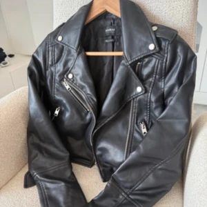 Svart skinnjacka från Pull&Bear - Säljer en svart skinnjacka från Pull&Bear.  Jackan har silvriga dragkedjor, bred krage och coola detaljer som axelklaffar och snedställda fickor. 