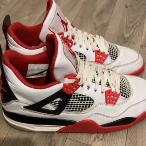 Nike Air Jordan 4 Fire Red sneakers - Nike Air Jordan 4 Fire Red sneakers med vit ovandel i skinn, röda och svarta detaljer samt meshpaneler på sidorna. Klassisk Jumpman-logga på plösen och röd sula. Skorna har snörning och en sportig, ikonisk design som sticker ut.