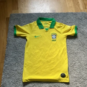 Brasilien fotbollströja Nike gul/grön - Säljer en klassisk Brasilien fotbollströja från Nike i gult med gröna detaljer på krage och ärmslut. Tröjan har korta ärmar, broderat landslagsemblem och Nike-logga på bröstet. Tillverkad i lätt och ventilerande material, perfekt för match eller träning.