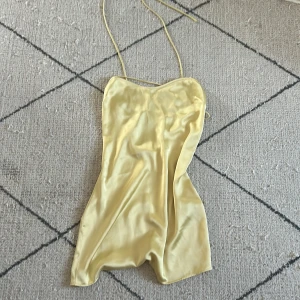 Gul satin kortklänning med smala band - Superfin ljusgul kortklänning i satin med tunna axelband och snygg detalj med korsade band över bysten. Klänningen har en lätt glansig finish och en enkel, figurnära passform som känns både trendig och fräsch.