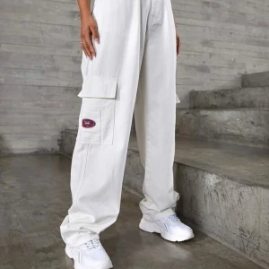 Vita wide cargopants med fickor - Säljer ett par vita cargopants med bred passform och stora fickor på sidorna. Byxorna har en cool patchdetalj i rött och blått på benet och bakfickan. Tillverkade i kraftig bomull, perfekt för en avslappnad streetstil.