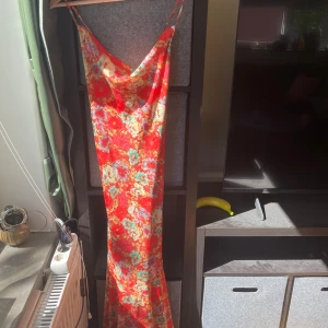 Adoore långklänning  - Supersnygg långklänning från Adoore i rött med blommigt mönster. Klänningen har tunna axelband, snörning i ryggen och en lätt glansig yta.