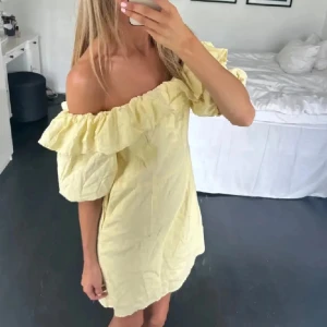 Gul offshoulder kortklänning med volang - Superfin ljusgul kortklänning med offshoulder och stora volanger upptill. Klänningen har en luftig passform och är perfekt för varma dagar. Snygg och trendig stil som passar dig som vill sticka ut lite extra.