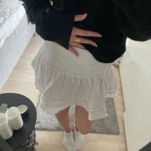 Vit volangkjol med smock - Supersöt vit kortkjol med smock i midjan och volangkant. Kjolen har broderade detaljer och är perfekt för varma dagar. Lätt och luftig känsla, passar till allt från sneakers till sandaler.