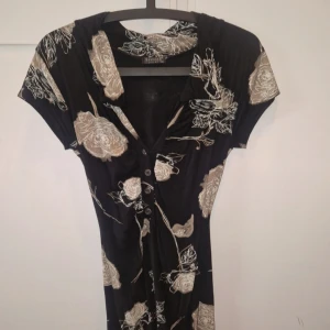 Svart blommig klänning  - Svart kortklänning från Sisters Point med stora beige blommor, v-ringning och knappar framtill. Klänningen har korta ärmar. Stl s/m