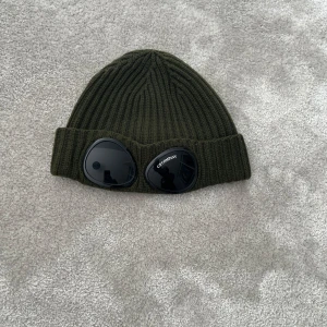 Olivgrön Goggle-mössa från C.P. Company - Säljer en olivgrön ribbstickad mössa från C.P. Company med ikoniska svarta goggle-linser framtill. Perfekt accessoar för dig som vill sticka ut och hålla dig varm. Bra skick och bara att höra av sig vid frågor!