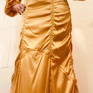 Guldig långklänning BY MALENE BIRGER - Snygg långklänning i guldig satin med markerad rynkning längs hela klänningen. Modellen har långa puffärmar och en figurnära passform som ger en lyxig och trendig look. Perfekt för dig som vill sticka ut med en elegant och modern stil. Aldrig använd. 