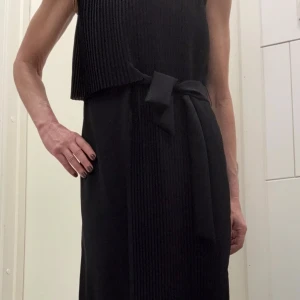 Svart plisserad detalj klänning med knytband - Snygg svart klänning med plisserad överdel och slät nederdel. Knytband i midjan ger en cool och stilren look. Ärmlös modell som passar perfekt för dig som gillar minimalistisk design och vill ha något enkelt men ändå unikt.
