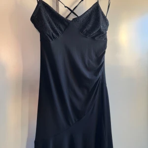 Svart långklänning från Glance - Elegant svart kort klänning från Glance med tunna axelband som korsas i ryggen. Överdelen har vackra broderier och pärlor som ger en lyxig känsla. Klänningen har en mjuk, följsam passform och är perfekt för dig som vill sticka ut med stilrena detaljer. Aldrig använt klänningen köptes secondhand! 