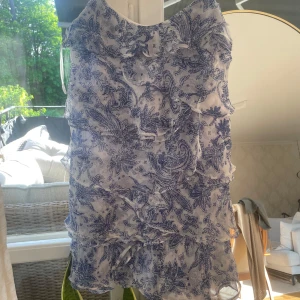 Blåvit blommig volangklänning - Supersöt klänning i  vitt med blått blommönster och flera volanglager. Ärmlös modell i lätt och luftig polyester, perfekt för varma dagar.