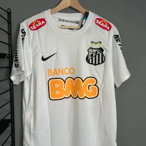 Vit Santos Neymar Jr fotbollströja Nike - Santos FC matchtröja i vitt med Neymar Jr och nummer 11 på ryggen. Tröjan har korta ärmar, svarta tryck och flera sponsorloggor i orange och svart. Tillverkad i lätt och ventilerande material, perfekt för fotbollsälskare. Helt ny med tagg en replika 