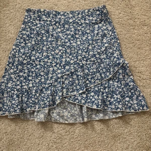 Blå blommig omlottkjol med volang - Supersöt blå kortkjol med vitt blommönster och volangkant. Kjolen har omlottmodell som ger en snygg och flowy känsla. Perfekt för dig som gillar en feminin och somrig stil. Lätt och skön att bära. Storlek 146 men passar mig som oftast bär storlek xs.💓