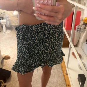 Grön blommig omlottkjol med volang - Supersöt grön kortkjol med vitt blommönster och volangkant. Kjolen har omlottmodell med knyt i sidan som ger en snygg och feminin look. Perfekt för varma dagar när du vill känna dig både chill och stylish. Storlek 140 men passar mig som oftast bär storlek xs!💓