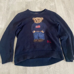 Mörkblå stickad tröja Polo Bear - Mörkblå stickad tröja från Polo Ralph Lauren med ikoniska Polo Bear-motivet framtill. Tröjan har rund hals, ribbade muddar och RL-broderi i rött. Perfekt för dig som gillar klassisk preppy stil med en twist.