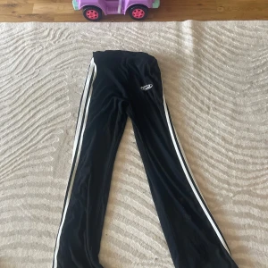 Svarta mjukisbyxor med vita ränder - Svarta mjukisbyxor med vita ränder längs sidorna och en liten vit logga på låret. Byxorna har en relaxed passform och är perfekta för chill dagar eller träning. Mjukt material som känns skönt mot huden.