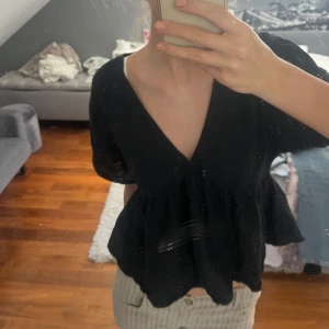 Svart v-ringad blus med volang - Säljer en svart blus med djup v-ringning och volang nertill. Modellen är croppad och har korta, lösa ärmar som ger en avslappnad vibe. Bara använd 1-2 gånger 🤩