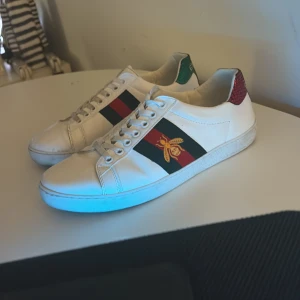 Gucci ace 40 - Gucci Ace sneakers i vitt skinn med klassiska gröna och röda ränder på sidorna. Ena sidan har en broderad guldfärgad bi. Baksidan har röd och grön hälflik i ormskinnslook. Snygga och ikoniska sneakers med snörning och rund tå.