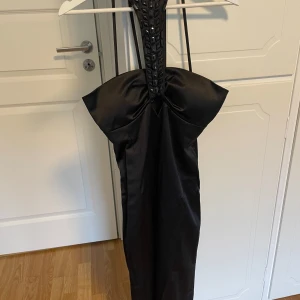 Svart långklänning från Fionella - Supersnygg svart långklänning från Fionella med unik halterneck och bred dekorerad axelrem. Klänningen har en stor rosett framtill och är figurnära med dragkedja i ryggen. Perfekt för dig som vill sticka ut med en elegant och cool look.