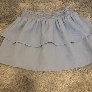 Ljusblå volangkjol  - Supersöt ljusblå kortkjol med volang och smock i midjan. Kjolen är i ett lätt och luftigt tyg, perfekt för varma dagar. Enkel att matcha med både t-shirt och linne för en chill look.