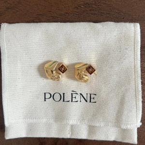 Polène Eroz örhängen i 24k guld - Nypris 140€! Säljer ett par Polène Eroz stud earrings, tillverkade i 24 karats guldplätering. Örhängena har en unik, skulptural form med både blanka och råa ytor samt en liten fyrkantig detalj med Polène-logga. Perfekta för dig som gillar statement-smycken med lyxig känsla.