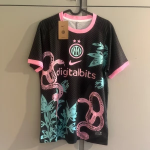Inter Milan x Nike svart fotbollströja - Inter Milan fotbollströja från Nike i svart med rosa och turkosa detaljer. Tröjan har korta ärmar, rosa ormmönster och turkosa blad, samt klubbmärke och sponsortryck på bröstet. Tillverkad i lätt och ventilerande material, perfekt för match eller träning.