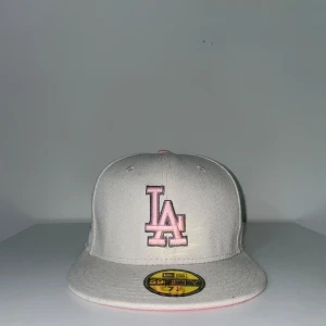 Grå och rosa LA-keps från New Era - Snygg grå keps från New Era med LA-logga i rosa framtill och broderad D med blommor på sidan. Kepsen har rosa detaljer och klassisk 59FIFTY-form. Perfekt för dig som vill sticka ut med en stilren och unik accessoar.