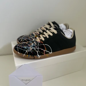 Maison Margiela sneakers med färgstänk - Unika svarta sneakers från Maison Margiela med coola färgstänk i blått, vitt, rött och gult över hela ovandelen. Skorna har beige snörning och bruna gummisulor. Perfekt för dig som vill sticka ut med en konstnärlig vibe och streetstyle-känsla.