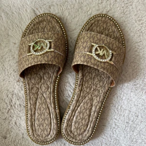 Beige sandaler Michael Kors strl 36 - Snygga beige sandaler från Michael Kors med MK-logga i guld och strass framtill. Skorna har mönstrad ovandel med logotyptryck och dekorativa stenar runt sulan. Perfekta för sommaren och ger en lyxig touch till din outfit. Oanvända. Fick dessa i present så vet inte om de är äkta 