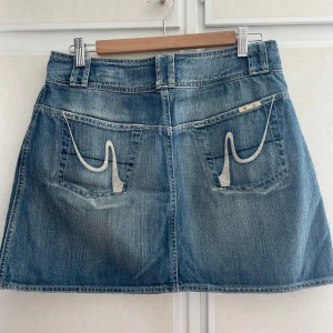 Blå vintage jeanskjol från Zoul,  - Snygg blå jeanskjol från Zoul i klassisk modell med dubbla knappar framtill, fickor både fram och bak samt coola kontrastsömmar. Kjolen har en lätt A-linjeform och broderade detaljer på bakfickorna. Storlek 42 men passar 36/38