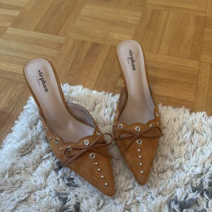 Bruna mocka mules från Stephan Paris - Snygga bruna mules i mocka med spetsig tå, låg klack och dekorativa silverfärgade nitar. Skorna har en söt rosett framtill och vågig kant längs öppningen. Perfekta för dig som vill ha en trendig och chic look.