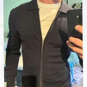 Mörkblå stickad kofta från J.Lindeberg - Snygg mörkblå stickad kofta från J.Lindeberg med dragkedja framtill och klassisk krage. Tröjan har en bröstficka och långa ärmar med ribbade muddar. Perfekt för lager-på-lager och stilren look.