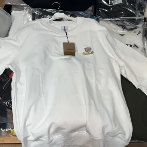 Vit sweatshirt från Burberry med emblem - Vit sweatshirt från Burberry med broderat emblem på bröstet i guld, svart och rött. Klassisk rund halsringning och långärmad modell. Mjuk bomullskvalitet som känns lyxig och stilren. Perfekt för dig som gillar exklusiva streetwear-vibbar. Finns i fler storlekar 